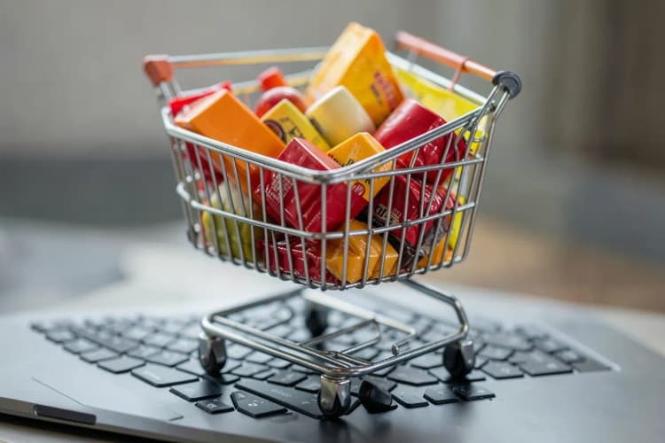 Um carrinho de compras de supermercado cheio de produtos, em cima de um teclado de computador.