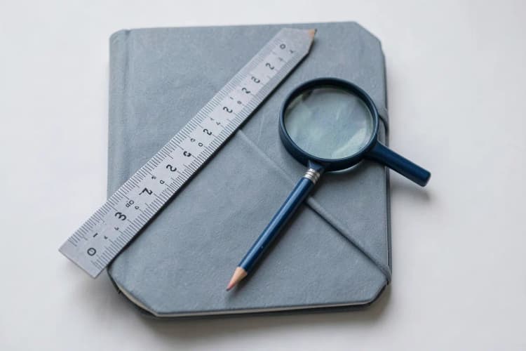 Ferramentas de escritório como régua, lápis e lupa sobre um caderno fechado, em tons de cinza e azul.